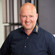 Maler-Busch-Maik - AGP MEDIA Smiling man with a shaved head wearing a dark blue button-down shirt in an office setting.