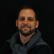 michael_waldinger - AGP MEDIA Portrait of a man with short dark hair and beard, wearing a gray scarf, smiling against a dark background.